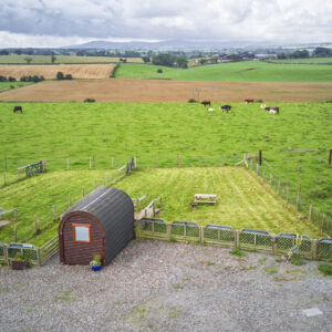 Aerial view of a single camping pod beside a fenced grass pitch, with open countryside and grazing fields in the distance.