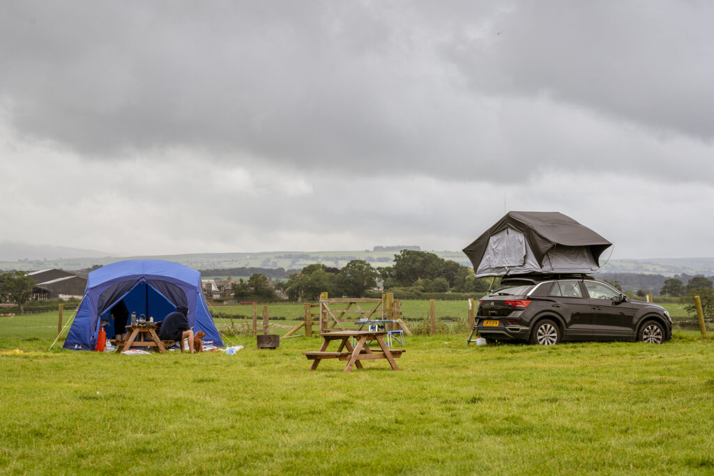 Blue family tent and a black car with a roof tent pitched on a grassy field at Camping at Cardewlees, with picnic benches and rolling countryside views under a cloudy sky.