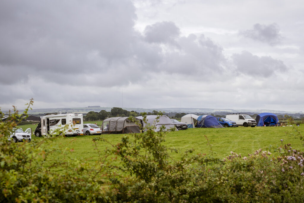 Row of tents, campervans and cars pitched along the edge of a grassy camping field at Camping at Cardewlees, with countryside views and a cloudy sky.