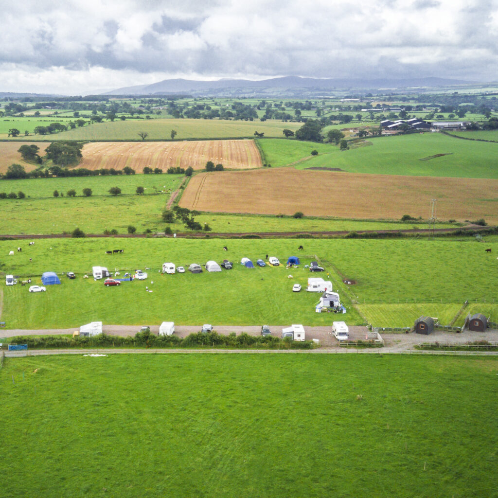 Aerial view of Camping at Cardewlees’ main field with tents and caravans, surrounded by patchwork Cumbrian farmland and distant hills.