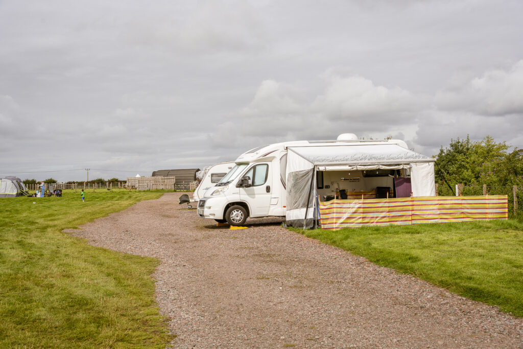 Motorhome parked on a gravel caravan pitch with a large awning and windbreak fencing, set on a grassy campsite under a cloudy sky.