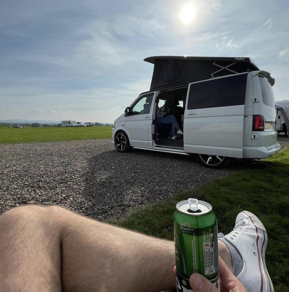 Campervan parked on a gravel pitch with the side door open and pop-top roof up, viewed from the grass with a drink can in the foreground under a sunny sky.
