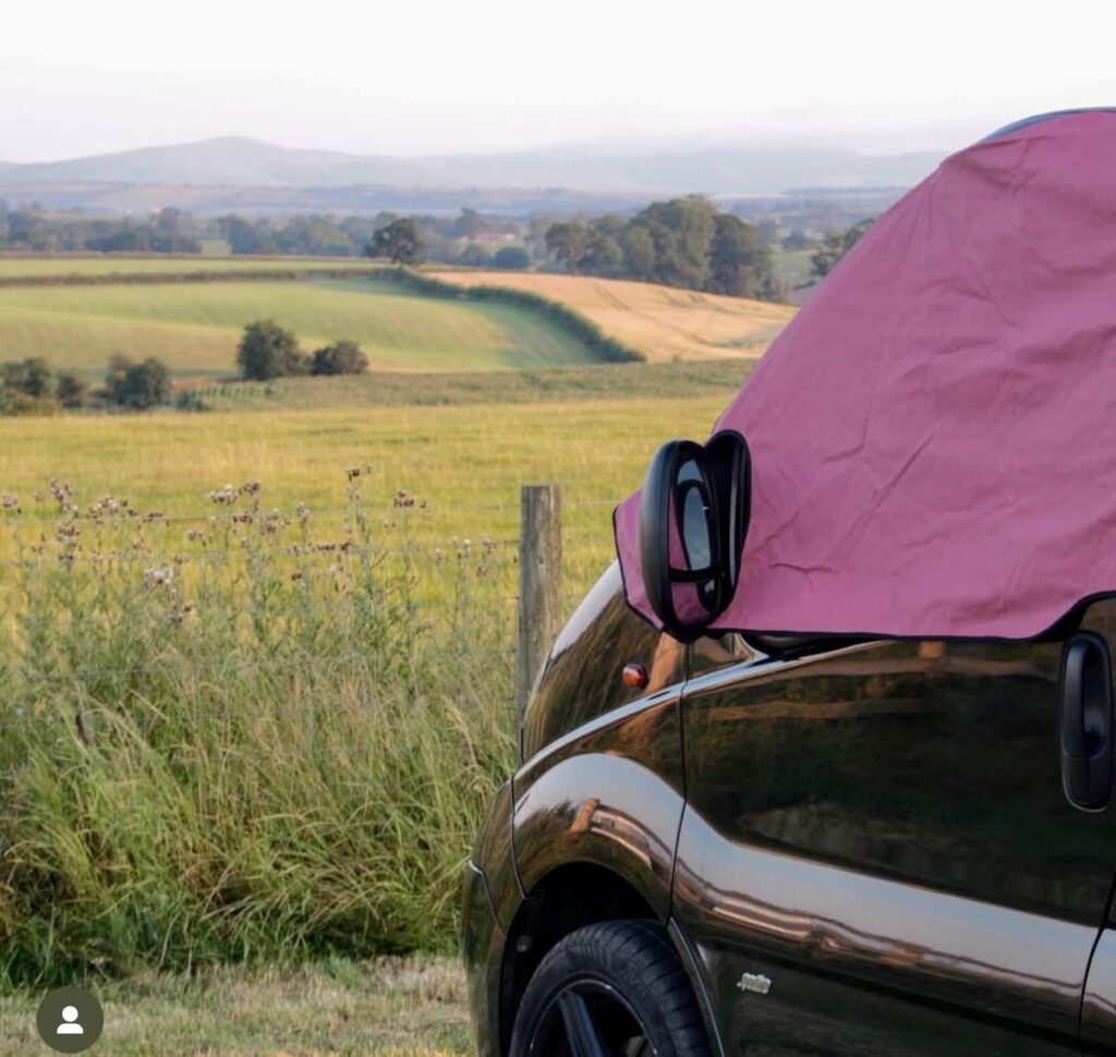 Car with a pink roof tent parked in a grassy field with wide countryside views and distant hills in the background.