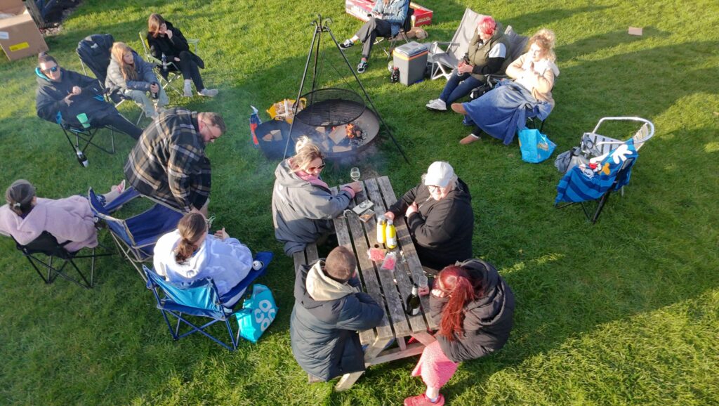 Overhead view of a group of campers sitting in folding chairs around a picnic table and a hanging fire pit on a grassy field, chatting and relaxing in the sun.