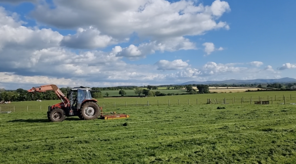 A tractor mowing a wide grassy field with open countryside views and a blue sky filled with clouds.