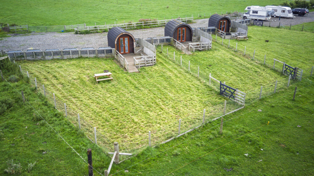 Aerial view of three camping pods with wooden decking and picnic benches, each beside a large fenced grass garden area.