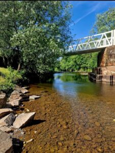 Shallow clear river with a pebbled shoreline and rocks, flowing under a white footbridge with trees along the banks.