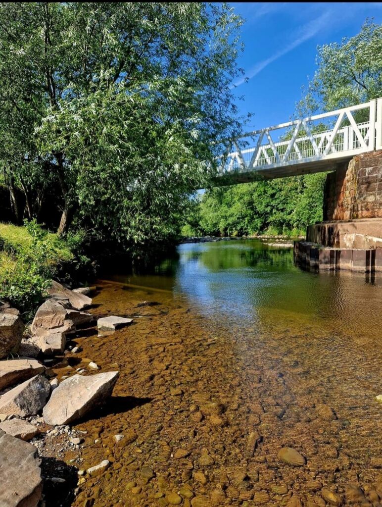 Shallow clear river with a pebbled shoreline and rocks, flowing under a white footbridge with trees along the banks.