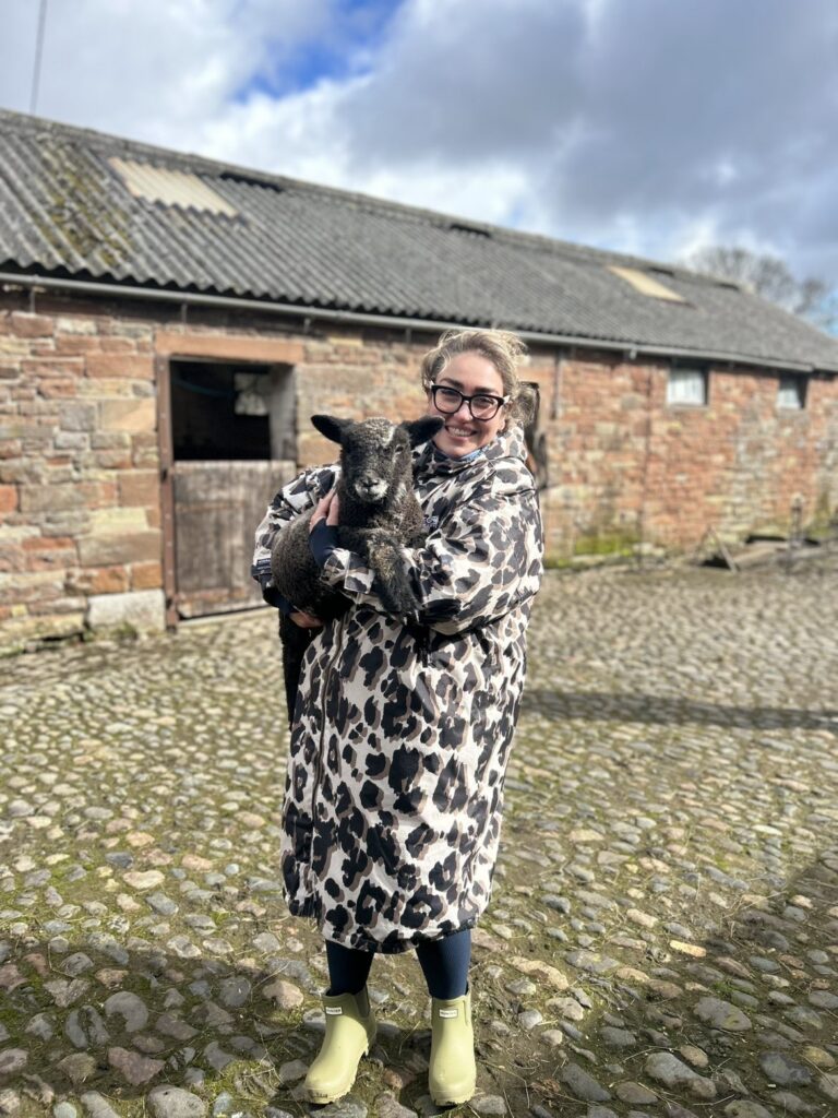 Woman in a patterned coat holding a small lamb in a cobbled farmyard outside a stone barn.