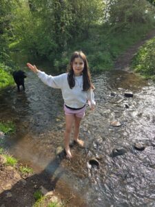 Child standing barefoot in a shallow woodland stream with one arm raised, while a black dog stands in the water nearby.