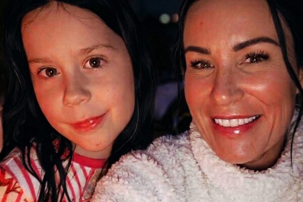 Close-up selfie of an adult and a child smiling at night, with warm string lights blurred in the background.