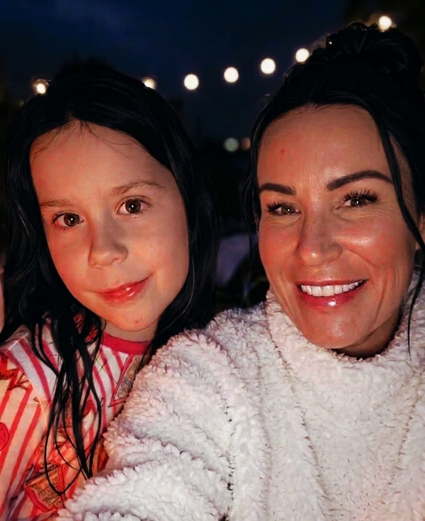Close-up selfie of an adult and a child smiling at night, with warm string lights blurred in the background.