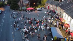 Aerial view of a busy community event in Dalston village square, with crowds gathered outside pubs and market stalls at dusk.
