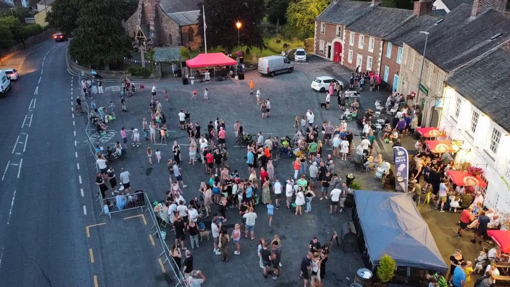Aerial view of a busy community event in Dalston village square, with crowds gathered outside pubs and market stalls at dusk.