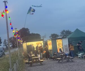 People gathered beside a brightly lit food trailer under colourful festoon lights at dusk.