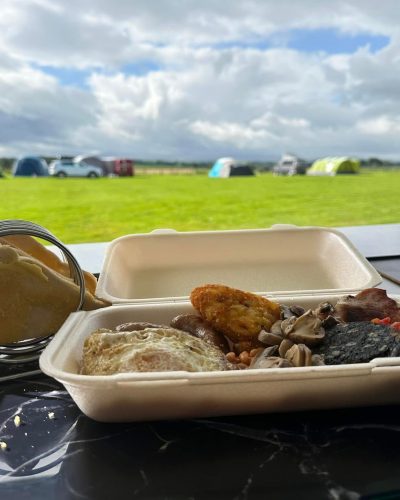 Takeaway full English breakfast in a compostable box on a counter, with bread rolls beside it and a campsite field with tents in the background.